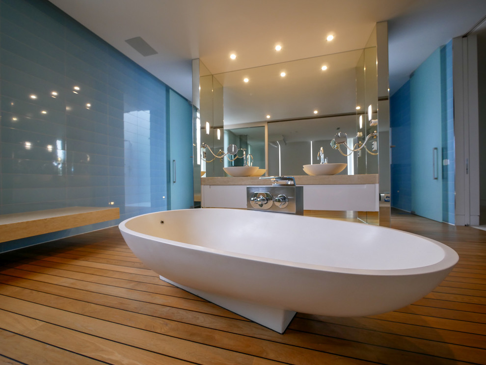 Large contemporary bathroom in Auckland with furniture-like cabinets, white cabinets, a freestanding tub, a wall-mount toilet, blue tile, subway tile, blue walls, light hardwood floors, a vessel sink, soapstone benchtops, brown floor and beige benchtops.