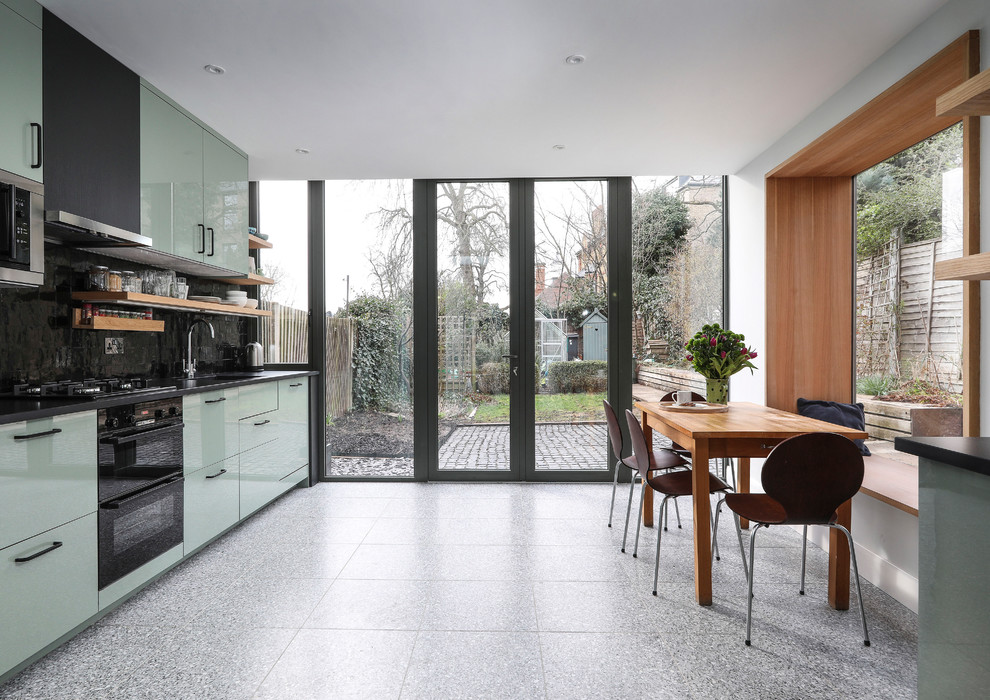 Photo of a mid-sized contemporary single-wall open plan kitchen in London with a drop-in sink, flat-panel cabinets, green cabinets, solid surface benchtops, black splashback, ceramic splashback, stainless steel appliances, terrazzo floors, grey floor and black benchtop.