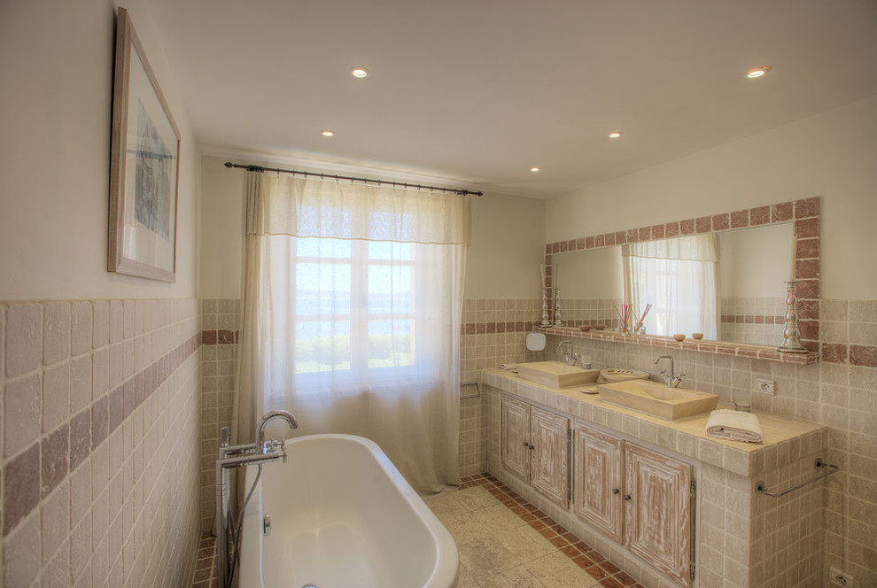 Photo of a mediterranean bathroom in Marseille with raised-panel cabinets, distressed cabinets, a freestanding tub, beige tile, red tile, beige walls, a vessel sink and beige floor.
