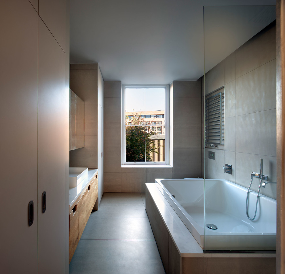 This is an example of a contemporary master bathroom in London with flat-panel cabinets, medium wood cabinets, a drop-in tub, a shower/bathtub combo, gray tile, grey walls, a vessel sink, grey floor, an open shower and white benchtops.