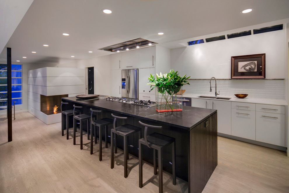 Photo of a large contemporary galley kitchen in Seattle with a double-bowl sink, flat-panel cabinets, white cabinets, solid surface benchtops, white splashback, glass tile splashback, stainless steel appliances, light hardwood floors and with island.