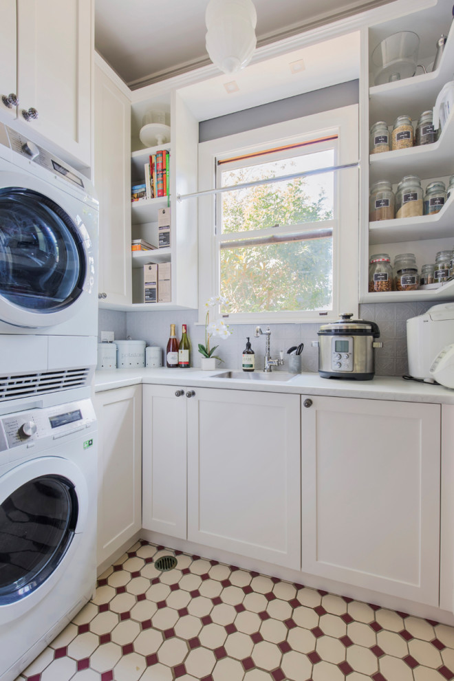 This is an example of a small traditional u-shaped utility room in Sydney with a single-bowl sink, shaker cabinets, white cabinets, quartz benchtops, grey walls, ceramic floors, a stacked washer and dryer, brown floor and white benchtop.