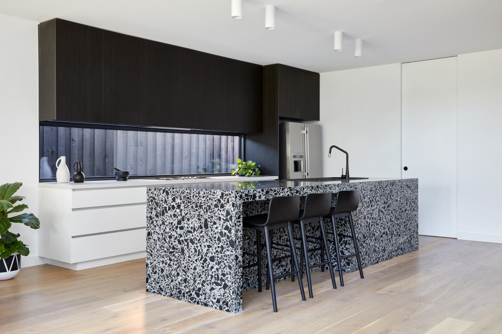 Large contemporary galley open plan kitchen with a double-bowl sink, flat-panel cabinets, black cabinets, terrazzo benchtops, window splashback, stainless steel appliances, light hardwood floors, with island and white benchtop.