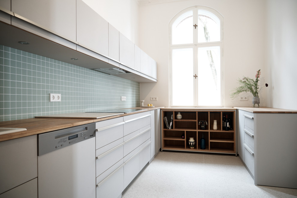 Design ideas for a modern kitchen in Berlin with flat-panel cabinets, grey cabinets, wood benchtops, green splashback, ceramic splashback, stainless steel appliances, terrazzo floors, no island, beige floor and brown benchtop.