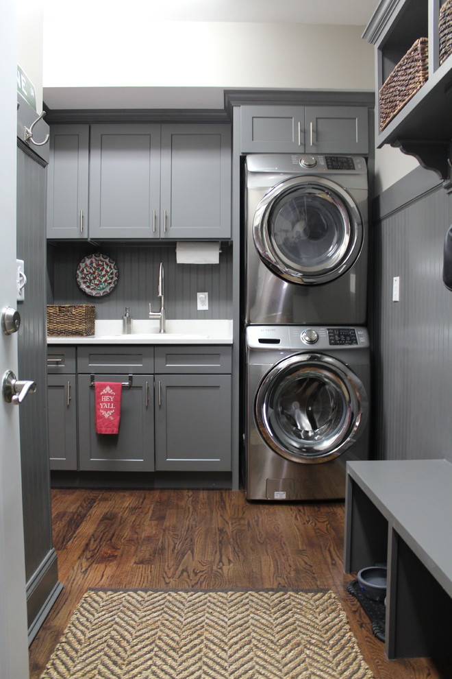 Photo of a mid-sized traditional single-wall dedicated laundry room in Raleigh with a drop-in sink, shaker cabinets, grey cabinets, white walls, dark hardwood floors and a stacked washer and dryer.