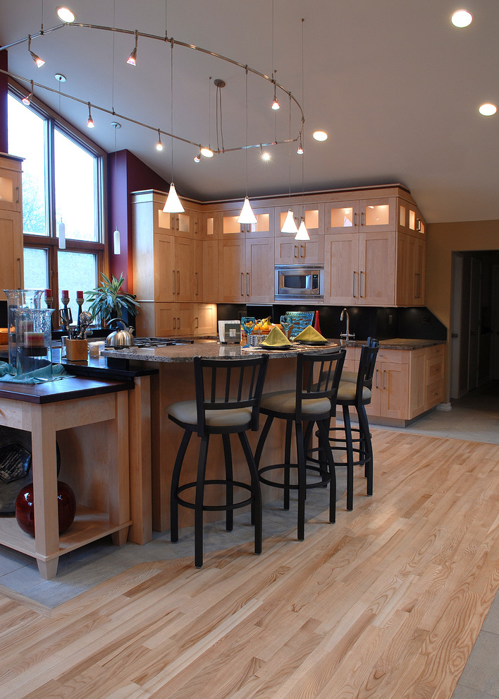 Photo of a mid-sized contemporary l-shaped eat-in kitchen in Chicago with an undermount sink, shaker cabinets, light wood cabinets, granite benchtops, black splashback, granite splashback, stainless steel appliances, light hardwood floors, with island, brown floor, multi-coloured benchtop and vaulted.