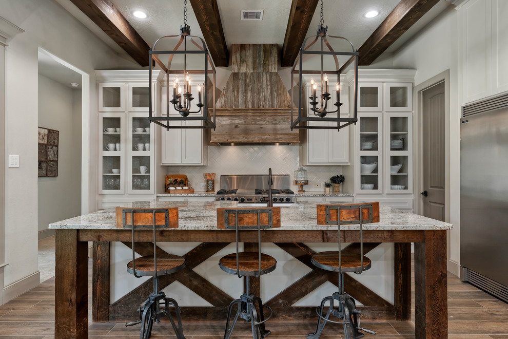 Mountain style l-shaped brown floor enclosed kitchen photo in Houston with a farmhouse sink, granite countertops, white backsplash, ceramic backsplash, stainless steel appliances, an island, glass-front cabinets, white cabinets and multicolored countertops