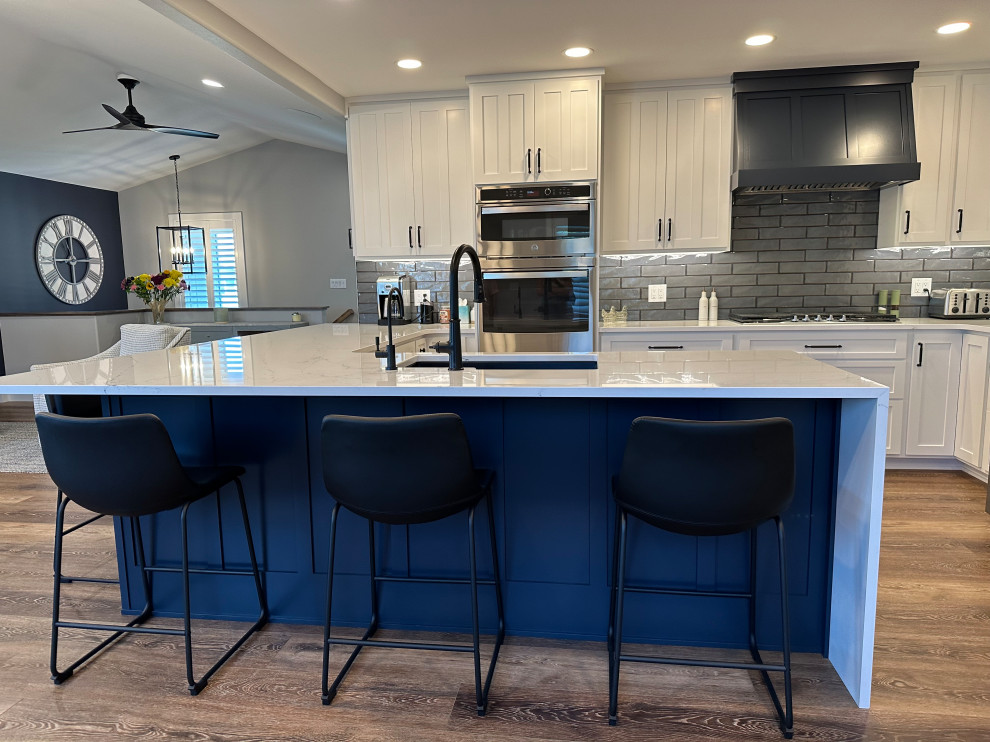 Example of a mid-sized transitional l-shaped medium tone wood floor and brown floor open concept kitchen design with an undermount sink, recessed-panel cabinets, white cabinets, quartz countertops, gray backsplash, ceramic backsplash, stainless steel appliances, an island and white countertops