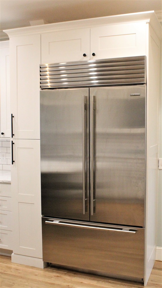 Large transitional galley light wood floor eat-in kitchen photo with a farmhouse sink, shaker cabinets, white cabinets, quartz countertops, white backsplash, ceramic backsplash, stainless steel appliances, an island and white countertops