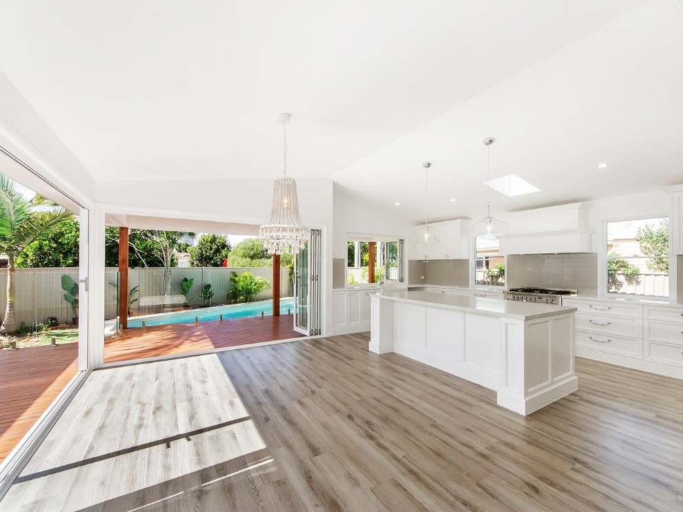 Photo of a large contemporary galley open plan kitchen in Gold Coast - Tweed with a farmhouse sink, shaker cabinets, white cabinets, quartz benchtops, beige splashback, subway tile splashback, stainless steel appliances, laminate floors, with island, grey floor and beige benchtop.