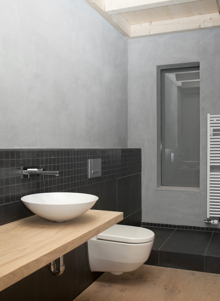 This is an example of an industrial bathroom in Berlin with open cabinets, a curbless shower, a wall-mount toilet, black tile, ceramic tile, grey walls, painted wood floors, a vessel sink and stainless steel benchtops.