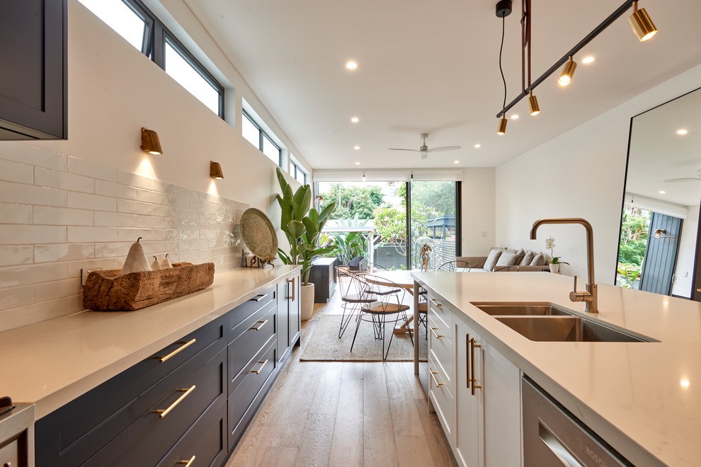 This is an example of an expansive beach style l-shaped open plan kitchen in Sydney with shaker cabinets, blue cabinets, quartz benchtops, subway tile splashback, with island, an undermount sink, white splashback, stainless steel appliances, light hardwood floors, brown floor and white benchtop.