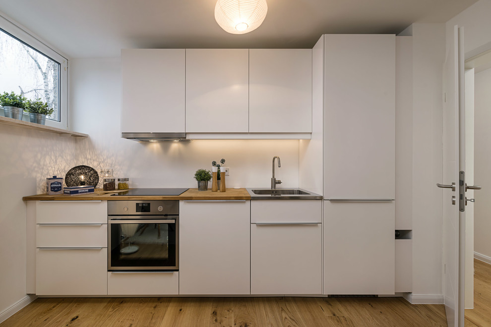 Offene, Einzeilige, Kleine Skandinavische Küche ohne Insel mit integriertem Waschbecken, flächenbündigen Schrankfronten, weißen Schränken, Arbeitsplatte aus Holz, Küchenrückwand in Weiß, Küchengeräten aus Edelstahl, braunem Holzboden, braunem Boden und brauner Arbeitsplatte in München