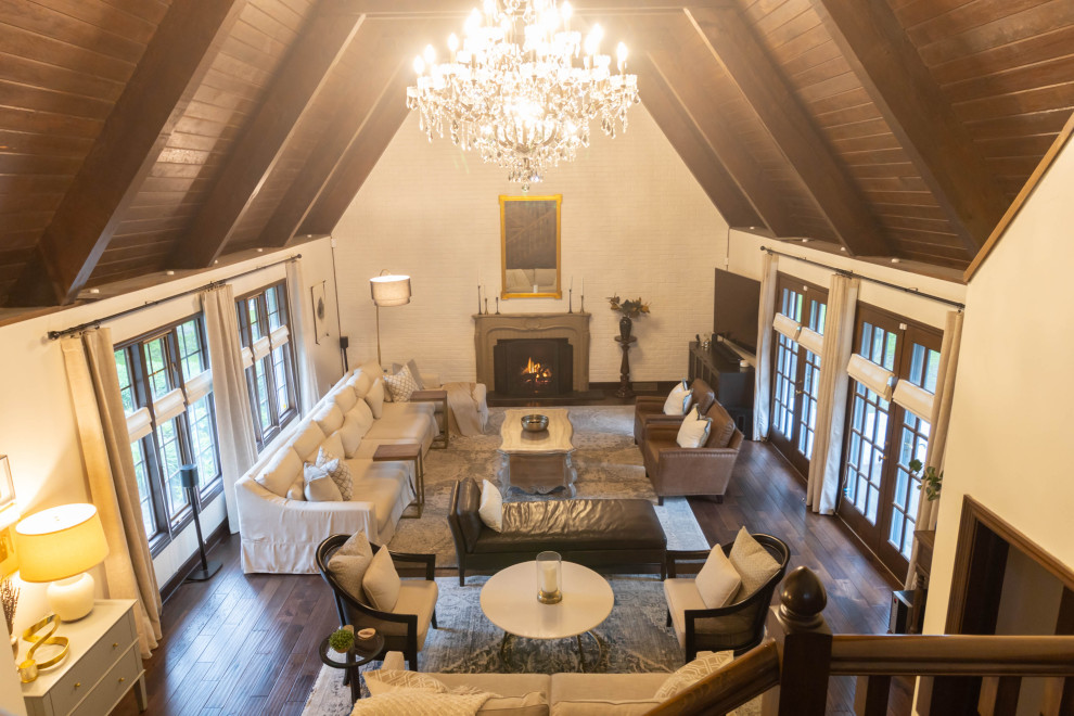 This is an example of a large classic formal mezzanine living room in Indianapolis with white walls, medium hardwood flooring, a standard fireplace, a brick fireplace surround, a corner tv, brown floors, exposed beams and brick walls.