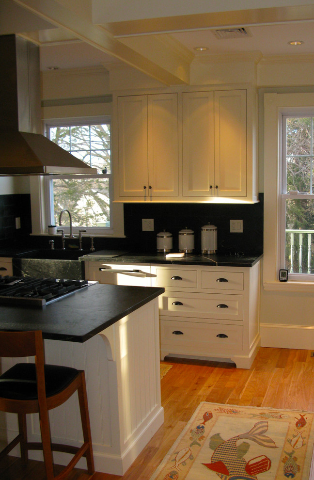 This is an example of a small traditional u-shaped kitchen pantry in Boston with shaker cabinets, white cabinets, a peninsula, a farmhouse sink, soapstone benchtops, black splashback, stainless steel appliances and light hardwood floors.