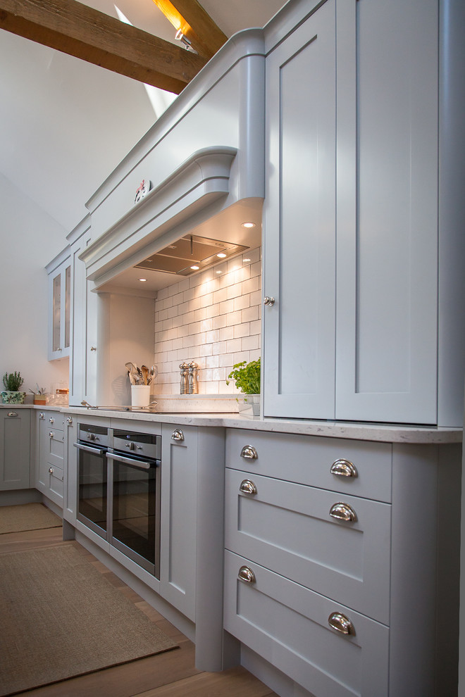 Mid-sized scandinavian l-shaped eat-in kitchen in Hertfordshire with a farmhouse sink, shaker cabinets, grey cabinets, quartz benchtops, white splashback, subway tile splashback, stainless steel appliances, medium hardwood floors, with island, brown floor and grey benchtop.