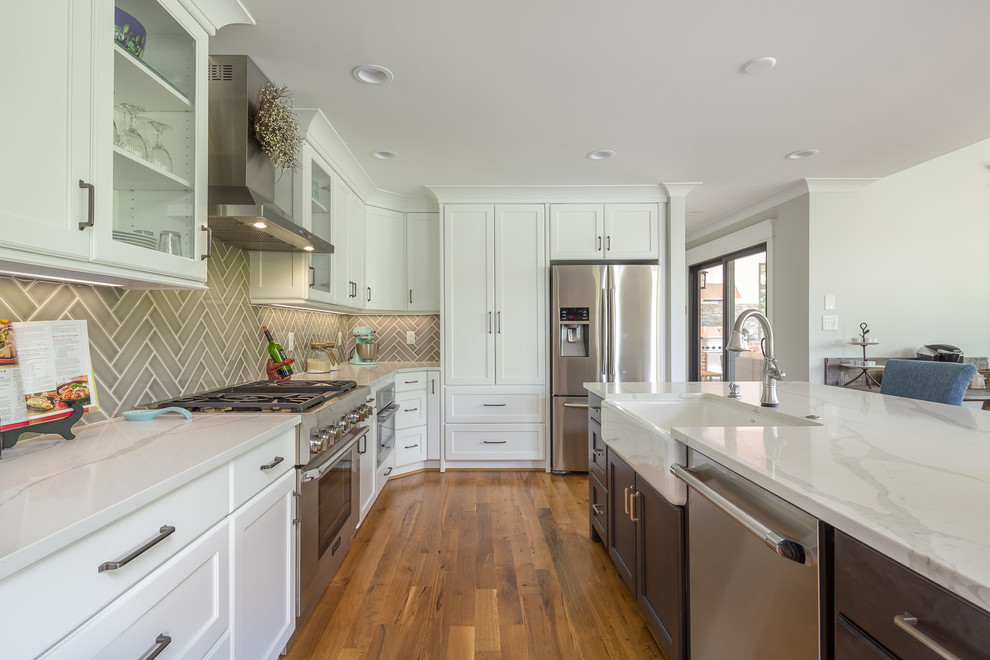 Photo of a mid-sized transitional l-shaped open plan kitchen in Raleigh with a farmhouse sink, shaker cabinets, white cabinets, quartz benchtops, grey splashback, ceramic splashback, stainless steel appliances, light hardwood floors, with island and white benchtop.