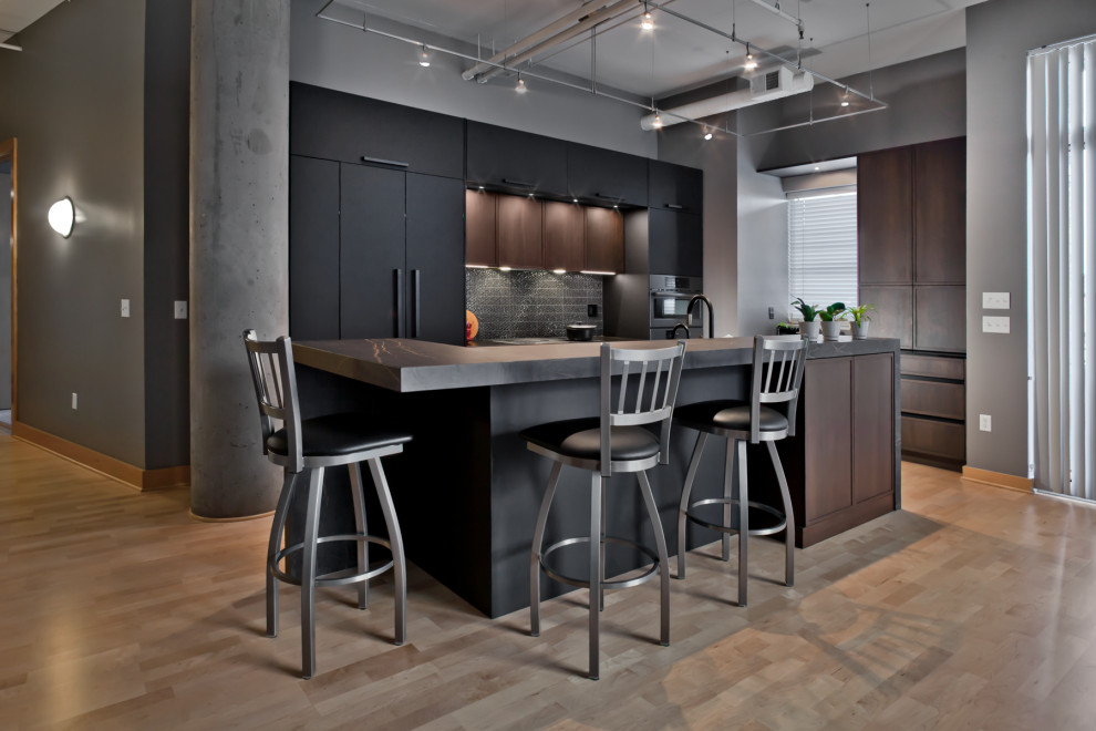 Photo of a medium sized industrial l-shaped kitchen/diner in Minneapolis with a submerged sink, black cabinets, engineered stone countertops, metallic splashback, porcelain splashback, integrated appliances, light hardwood flooring, an island, brown floors, black worktops and flat-panel cabinets.