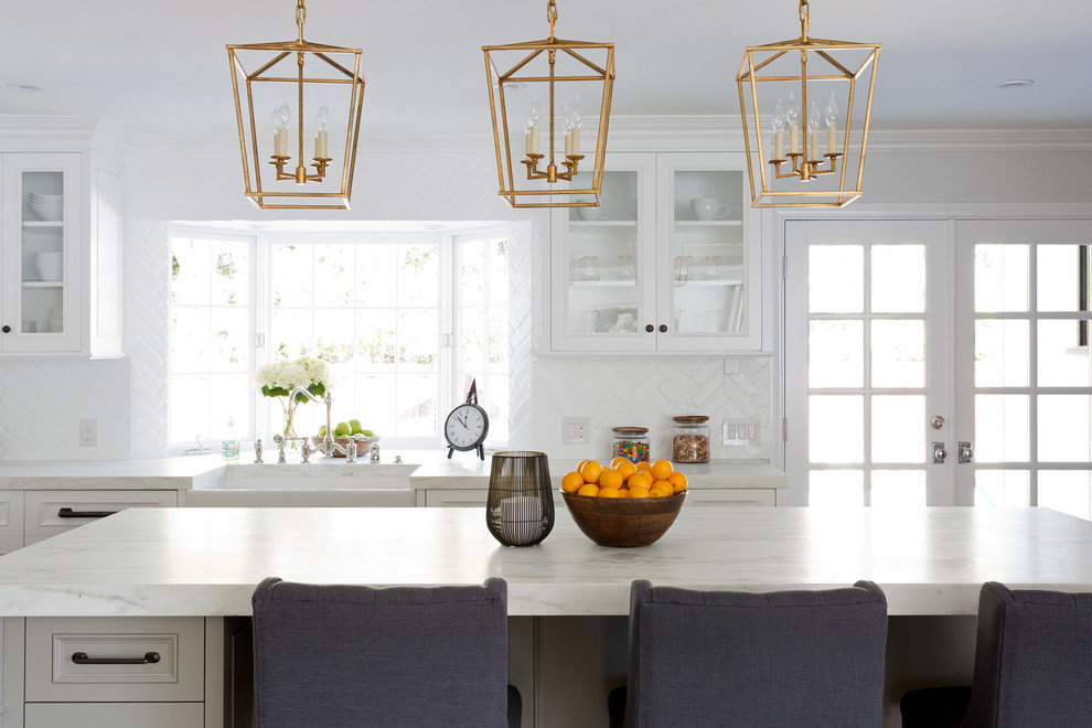Example of a large transitional l-shaped light wood floor open concept kitchen design in Orange County with a farmhouse sink, shaker cabinets, white cabinets, marble countertops, white backsplash, brick backsplash, stainless steel appliances and an island