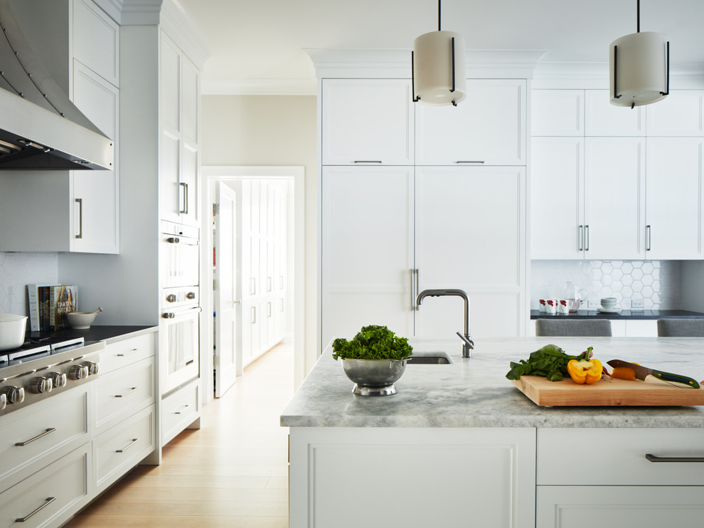 Photo of a large transitional u-shaped open plan kitchen in New York with an undermount sink, recessed-panel cabinets, white cabinets, solid surface benchtops, white splashback, mosaic tile splashback, stainless steel appliances, light hardwood floors, with island and black benchtop.