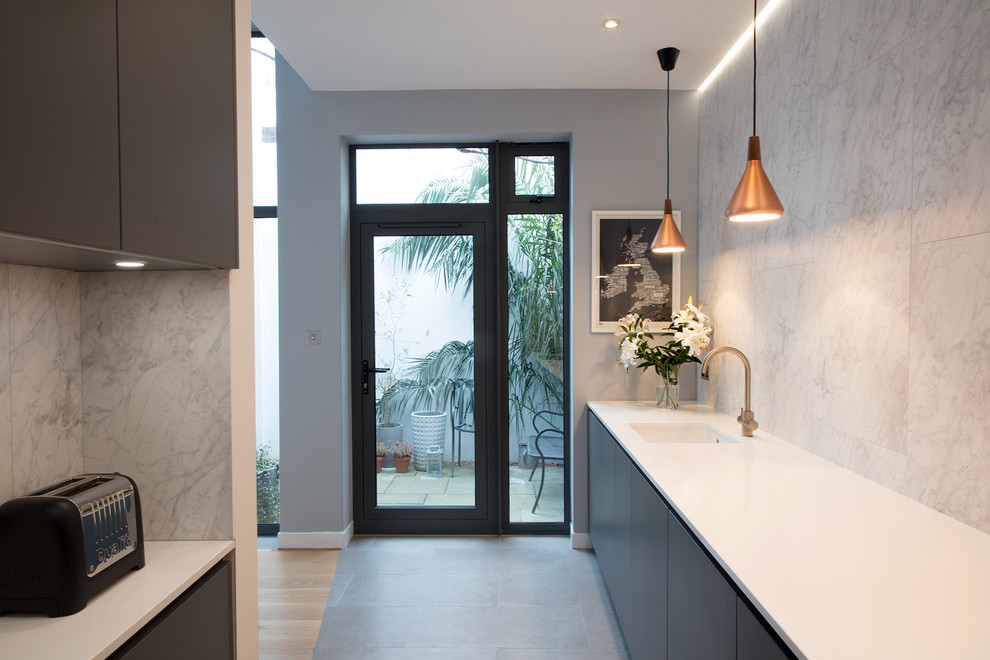 Design ideas for a large contemporary galley open plan kitchen in London with an integrated sink, flat-panel cabinets, grey cabinets, solid surface benchtops, white splashback, stone tile splashback, black appliances, concrete floors and grey floor.