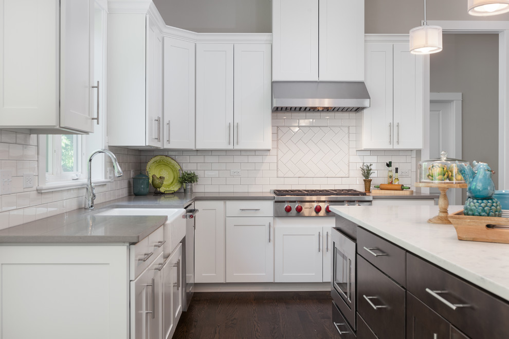 This is an example of a mid-sized transitional u-shaped open plan kitchen in Chicago with a farmhouse sink, shaker cabinets, white cabinets, white splashback, subway tile splashback, stainless steel appliances, dark hardwood floors, with island, brown floor, grey benchtop and quartz benchtops.