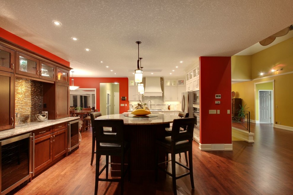 Open concept kitchen - large transitional l-shaped medium tone wood floor and brown floor open concept kitchen idea in Calgary with an undermount sink, recessed-panel cabinets, white cabinets, glass countertops, beige backsplash, matchstick tile backsplash, stainless steel appliances and an island