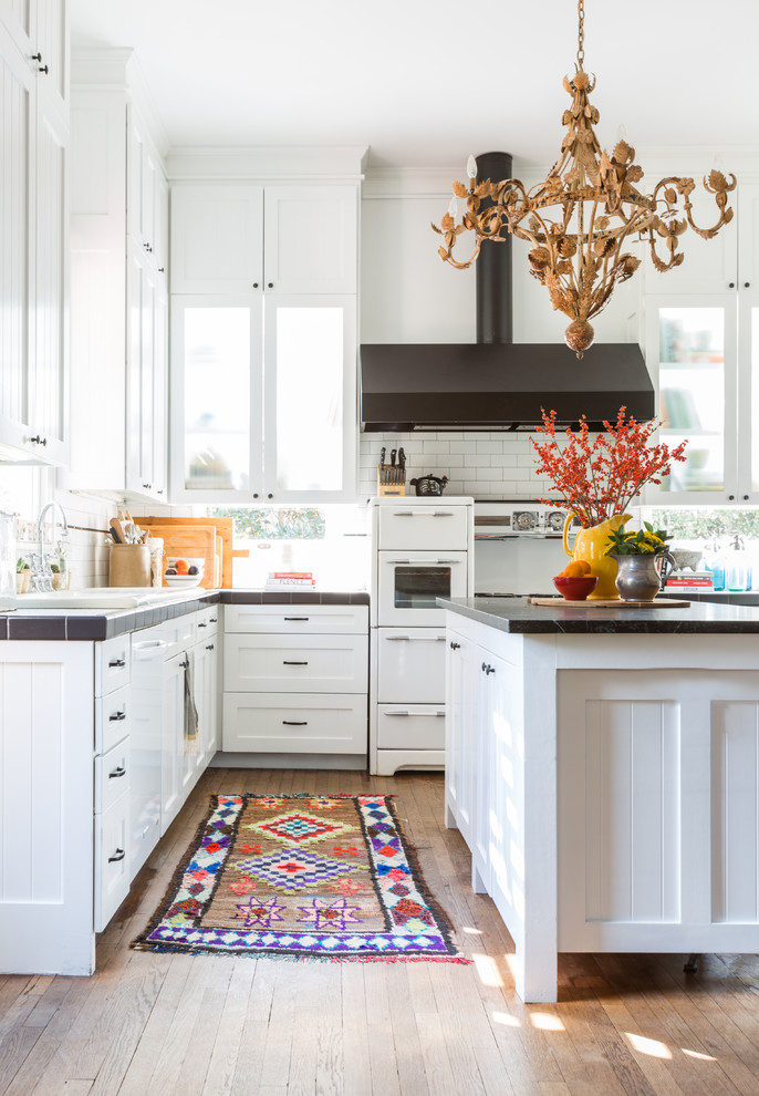 Design ideas for a mid-sized transitional u-shaped eat-in kitchen in Houston with shaker cabinets, white cabinets, tile benchtops, white splashback, subway tile splashback, white appliances, medium hardwood floors, with island, brown floor and a drop-in sink.