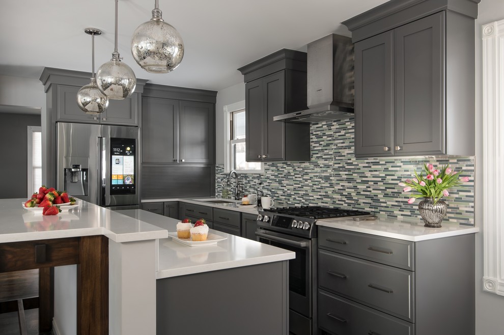 Inspiration for a mid-sized transitional l-shaped eat-in kitchen in Detroit with a drop-in sink, grey cabinets, multi-coloured splashback, stainless steel appliances, medium hardwood floors, with island, quartz benchtops, glass sheet splashback, recessed-panel cabinets, white benchtop and brown floor.