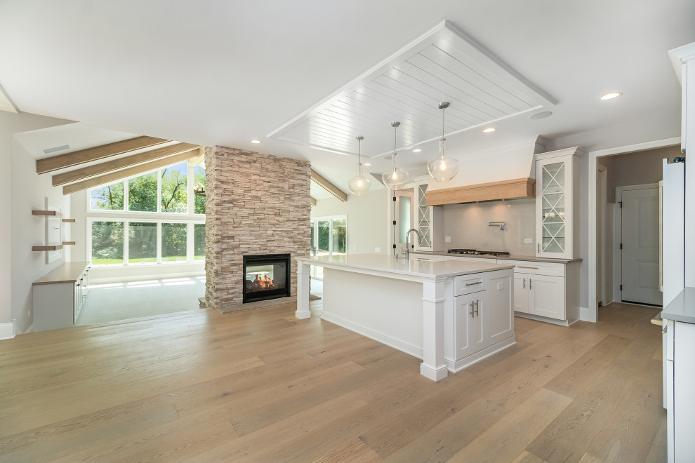 Large country l-shaped open plan kitchen in Chicago with a farmhouse sink, shaker cabinets, white cabinets, multi-coloured splashback, light hardwood floors, with island, beige floor, white benchtop and timber.