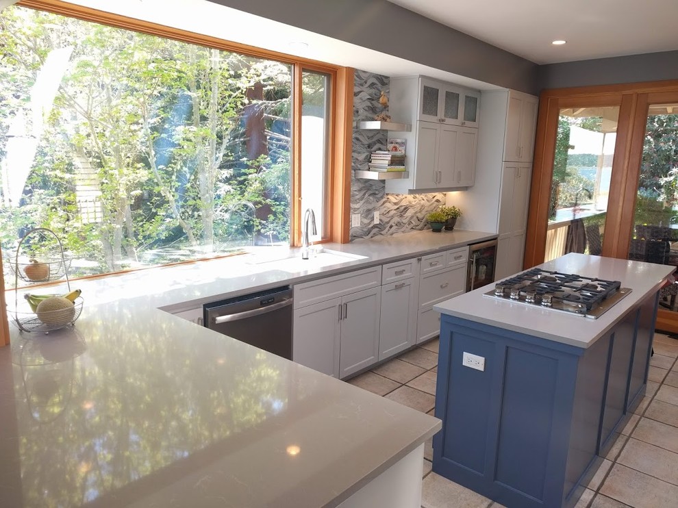 Photo of a mid-sized beach style l-shaped eat-in kitchen in Seattle with a double-bowl sink, flat-panel cabinets, grey cabinets, quartz benchtops, blue splashback, glass tile splashback, stainless steel appliances, ceramic floors, with island, grey floor and grey benchtop.