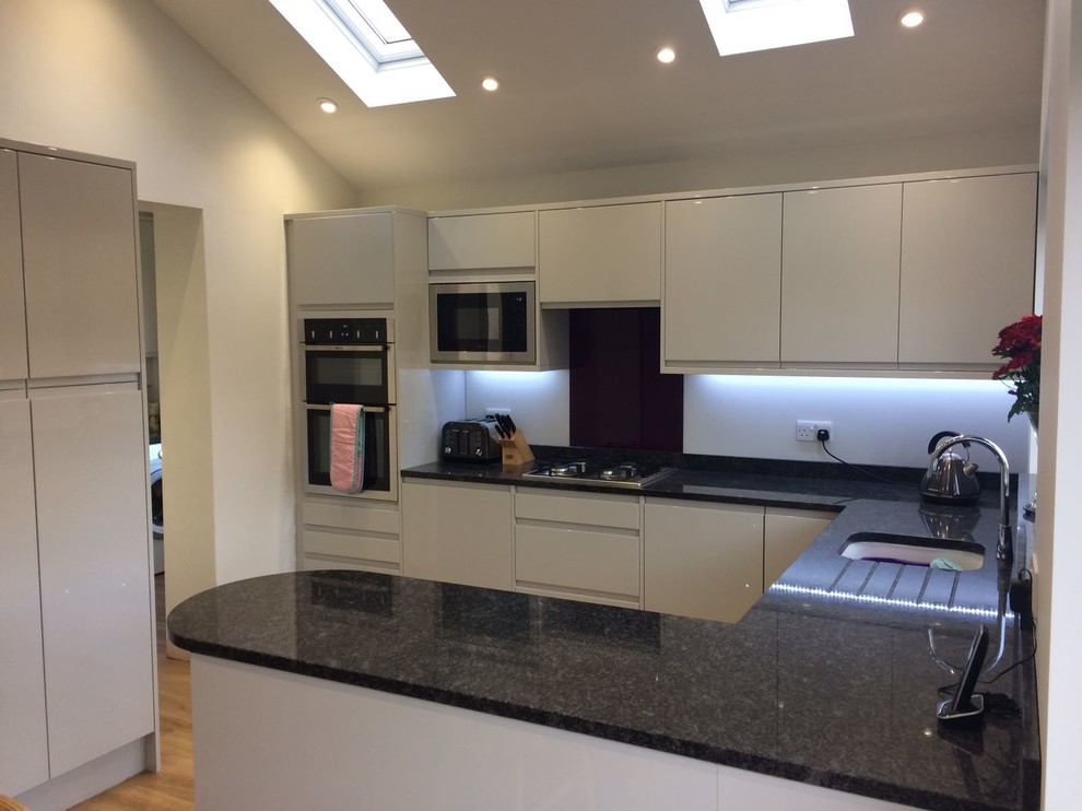 This is an example of a large contemporary u-shaped eat-in kitchen in Other with a drop-in sink, flat-panel cabinets, beige cabinets, granite benchtops, black splashback, glass sheet splashback, stainless steel appliances and no island.