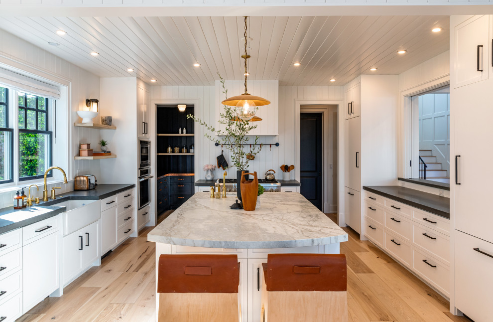 Design ideas for a large beach style eat-in kitchen in Los Angeles with a farmhouse sink, recessed-panel cabinets, white cabinets, concrete benchtops, white splashback, timber splashback, panelled appliances, light hardwood floors, with island, brown floor, grey benchtop and wood.
