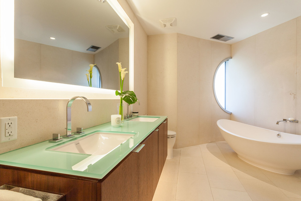 This is an example of a large modern master bathroom in Vancouver with flat-panel cabinets, medium wood cabinets, a freestanding tub, a one-piece toilet, beige walls, porcelain floors, an undermount sink, glass benchtops and beige floor.