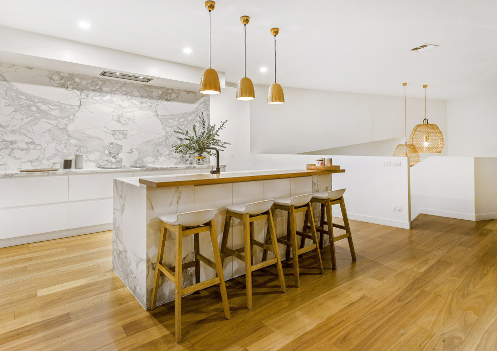 This is an example of a large coastal galley kitchen/diner in Melbourne with a submerged sink, white cabinets, marble worktops, grey splashback, marble splashback, black appliances, light hardwood flooring, multiple islands, grey worktops and a vaulted ceiling.