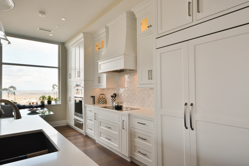 Example of a transitional single-wall medium tone wood floor and brown floor open concept kitchen design in Toronto with an undermount sink, shaker cabinets, white cabinets, quartz countertops, white backsplash, marble backsplash, paneled appliances, an island and white countertops