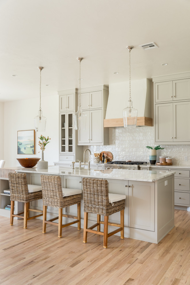 This is an example of a transitional galley kitchen in Oklahoma City with a farmhouse sink, shaker cabinets, grey cabinets, white splashback, stainless steel appliances, light hardwood floors, with island, beige floor and white benchtop.