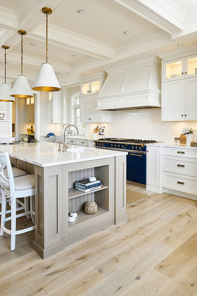 Inspiration for a large beach style l-shaped eat-in kitchen in Vancouver with a farmhouse sink, shaker cabinets, white cabinets, granite benchtops, white splashback, ceramic splashback, coloured appliances, light hardwood floors, with island, beige floor and white benchtop.
