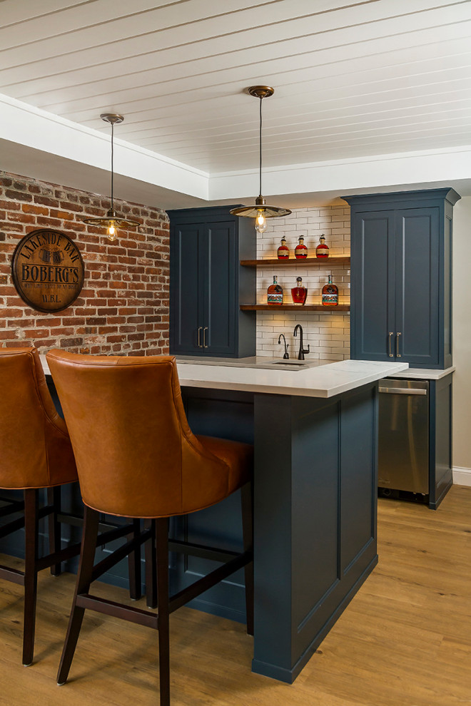 Photo of a mid-sized traditional galley seated home bar in Minneapolis with shaker cabinets, blue cabinets, white splashback, subway tile splashback, an undermount sink, brown floor and white benchtop.
