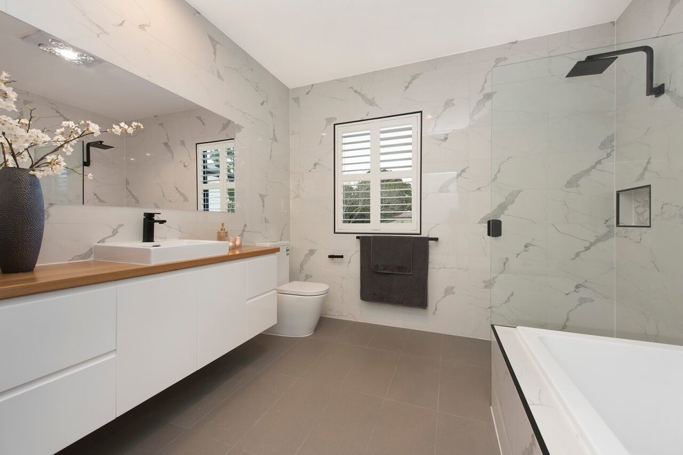 Photo of a contemporary master bathroom in Brisbane with flat-panel cabinets, white cabinets, a drop-in tub, a corner shower, a one-piece toilet, gray tile, white tile, grey walls, a vessel sink, wood benchtops, grey floor, an open shower and brown benchtops.