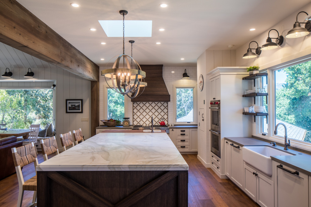 Design ideas for a country l-shaped open plan kitchen in Other with a farmhouse sink, shaker cabinets, white cabinets, white splashback, subway tile splashback, stainless steel appliances, dark hardwood floors, with island, brown floor and grey benchtop.