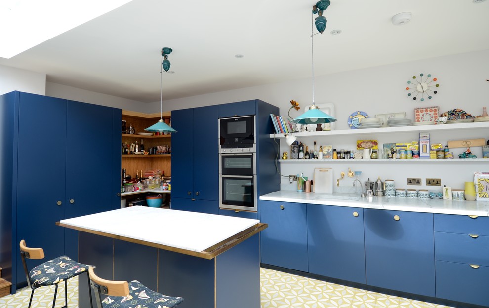This is an example of a mid-sized contemporary galley open plan kitchen in London with an integrated sink, flat-panel cabinets, blue cabinets, marble benchtops, white splashback, stone slab splashback, stainless steel appliances, travertine floors and with island.