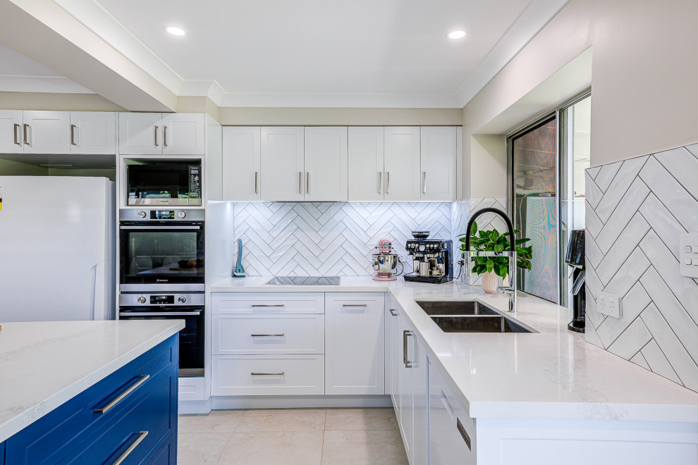 This is an example of a large contemporary u-shaped kitchen pantry in Brisbane with a double-bowl sink, shaker cabinets, white cabinets, quartz benchtops, white splashback, subway tile splashback, stainless steel appliances, ceramic floors, with island, beige floor and white benchtop.