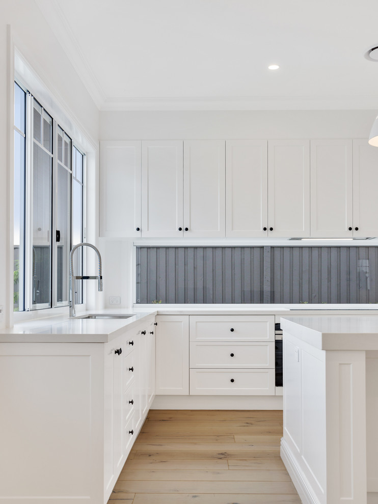 Inspiration for a beach style l-shaped kitchen in Brisbane with an undermount sink, shaker cabinets, white cabinets, window splashback, light hardwood floors, with island, brown floor and white benchtop.