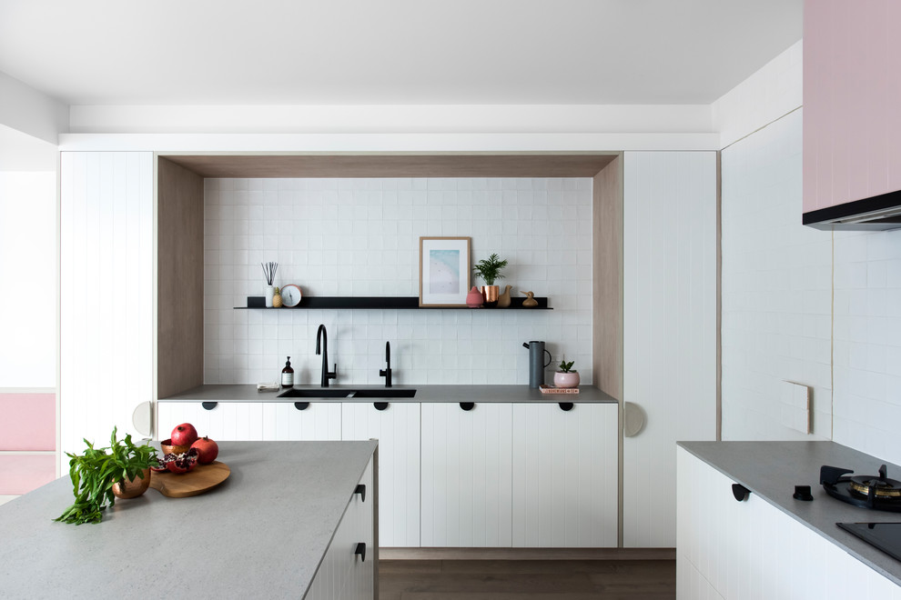 Mid-sized contemporary l-shaped open plan kitchen in Sydney with a double-bowl sink, white cabinets, recycled glass benchtops, white splashback, cement tile splashback, black appliances, medium hardwood floors, with island and grey benchtop.