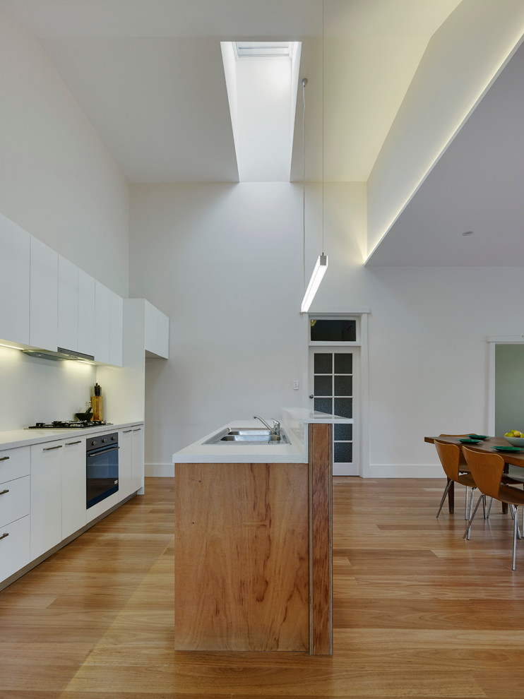 Inspiration for an expansive modern galley open plan kitchen in Sydney with a double-bowl sink, flat-panel cabinets, white cabinets, solid surface benchtops, white splashback, stone slab splashback, stainless steel appliances, light hardwood floors and with island.