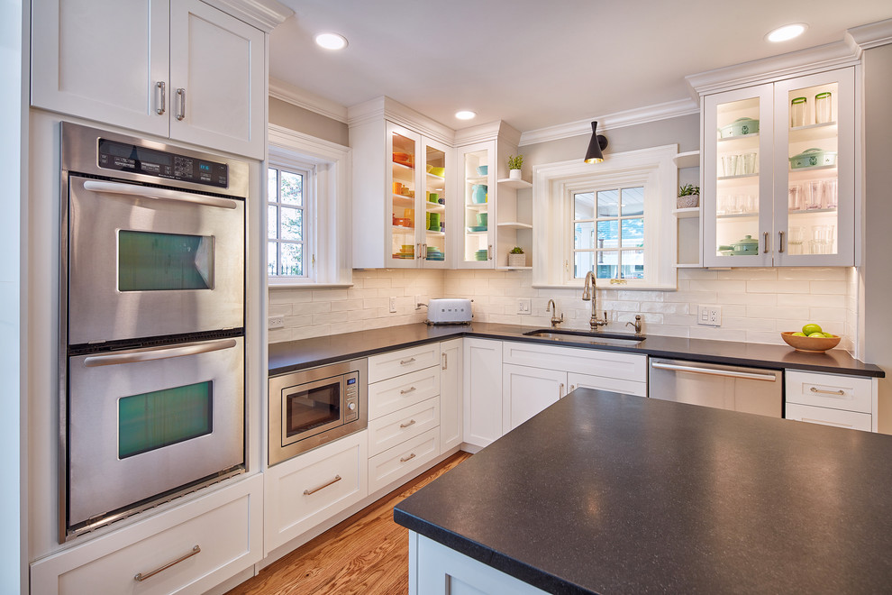 This is an example of a large transitional l-shaped kitchen in Kansas City with a double-bowl sink, white cabinets, granite benchtops, stainless steel appliances, with island, black benchtop, shaker cabinets, white splashback, subway tile splashback, medium hardwood floors and brown floor.