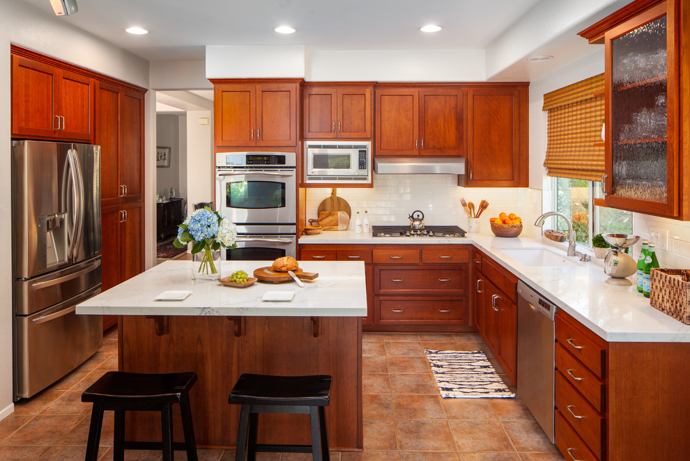 This is an example of a mid-sized transitional l-shaped eat-in kitchen in San Diego with an undermount sink, shaker cabinets, medium wood cabinets, quartz benchtops, white splashback, subway tile splashback, stainless steel appliances, porcelain floors, with island, multi-coloured floor and white benchtop.