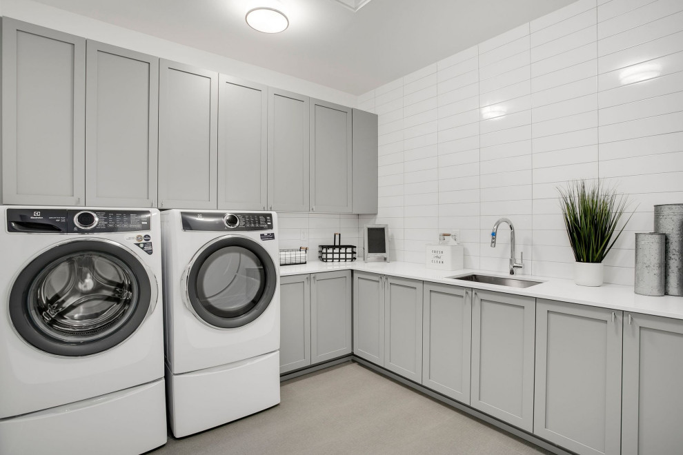 Modern inredning av en stor vita l-formad vitt tvättstuga enbart för tvätt, med en allbänk, luckor med infälld panel, grå skåp, bänkskiva i kvarts, vitt stänkskydd, stänkskydd i tunnelbanekakel, vita väggar, betonggolv, en tvättmaskin och torktumlare bredvid varandra och grått golv