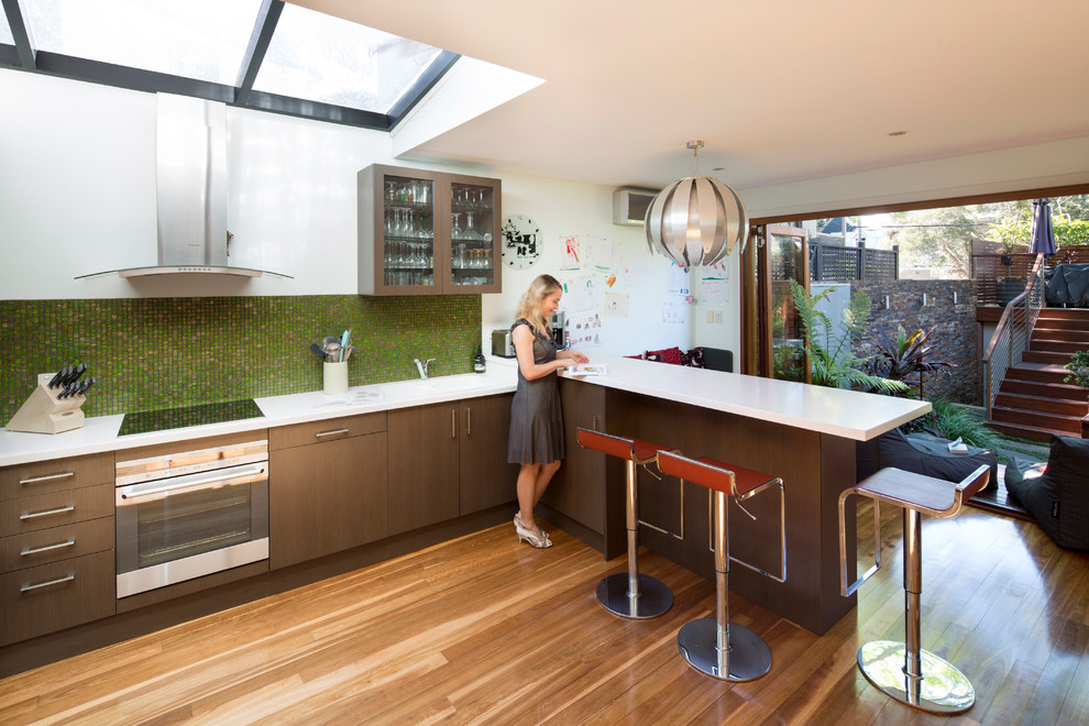 Contemporary l-shaped kitchen in Sydney with flat-panel cabinets, brown cabinets, green splashback, mosaic tile splashback, stainless steel appliances, light hardwood floors, a peninsula and beige floor.